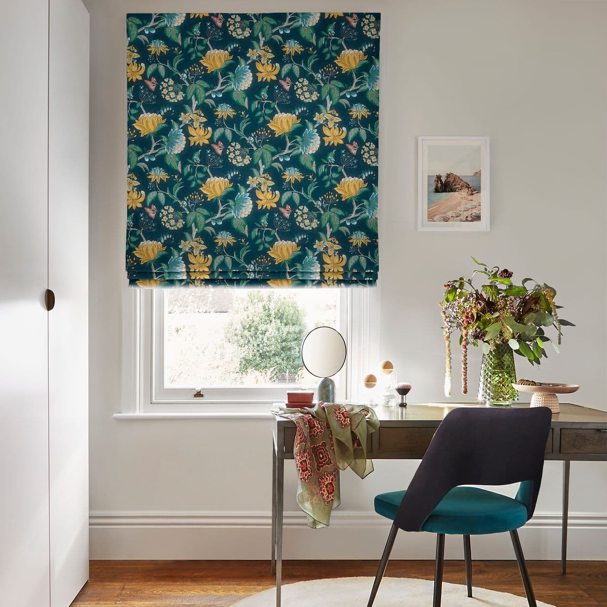Floral Roman blind with teal background and yellow flowers, lowered over a sash window, partially filtering daylight; small home office desk beneath with mirror, vase of foliage, teal chair, walls.
