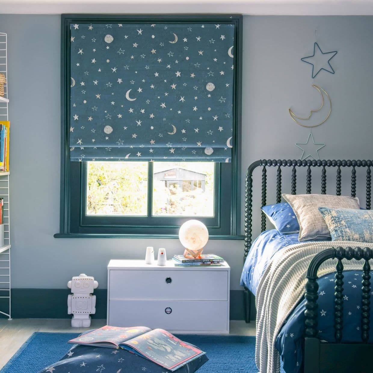 Navy Roman blind with moons and stars covering the window; lowered and filtering daylight; in a softly lit child's bedroom with starry bedding, bedside chest and hanging moon-star decor.