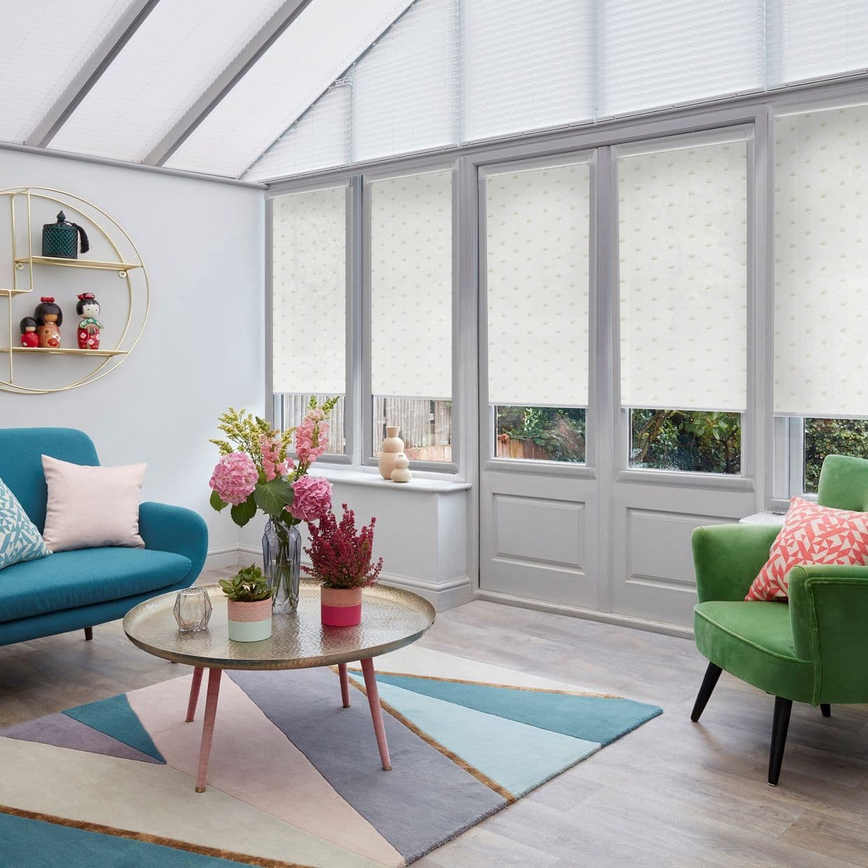 White patterned roller blinds, lowered over tall conservatory windows and doorâ€”covering upper panes while leaving lower glass visible; diffusing daylight in a bright sunroom with colorful sofas, rug, and flowers.