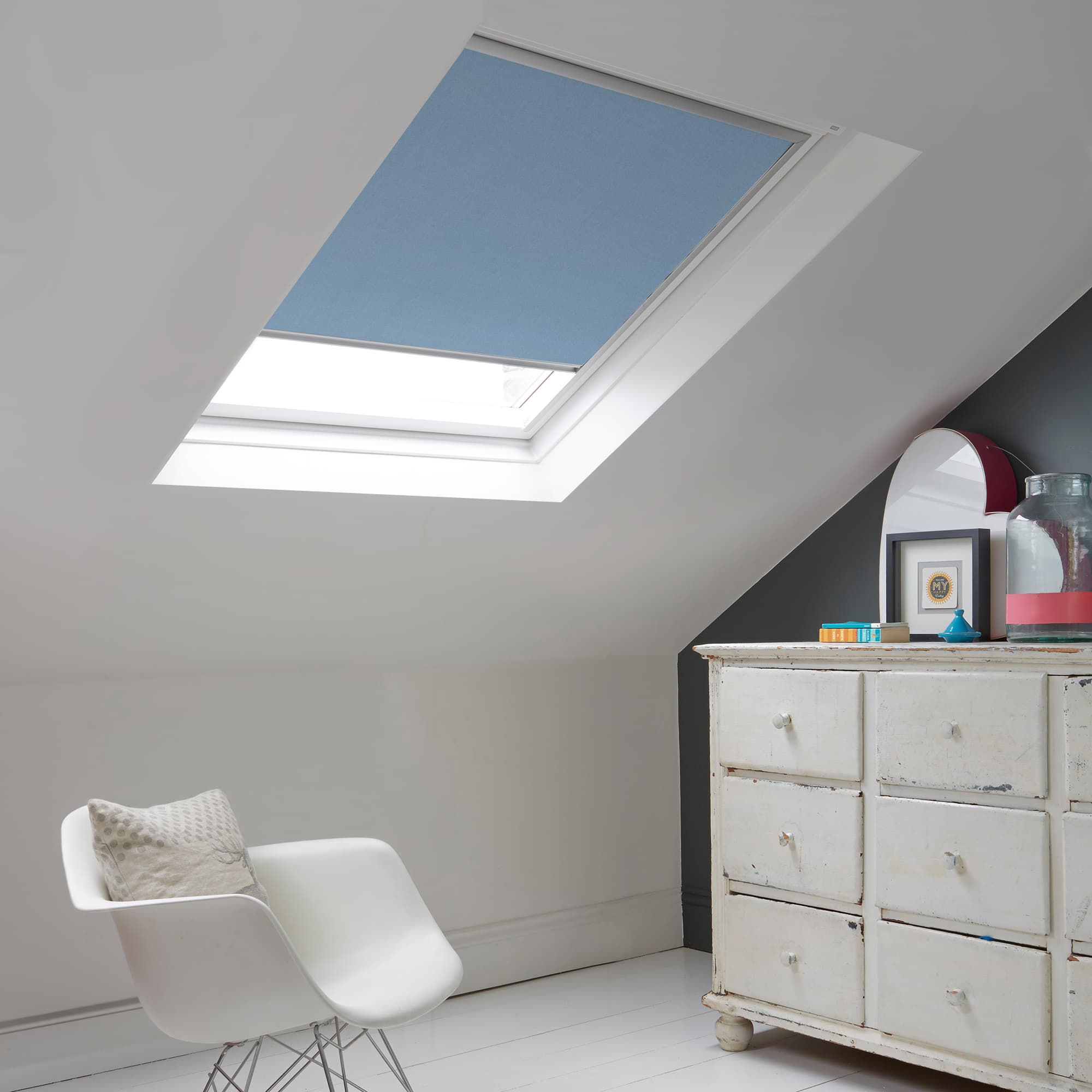 Room with white eaves wall, dark blue-grey wall to right with a shabby chic white drawer unit, a contemporary white chair, white painted floorboards and a skylight blind in mid blue.