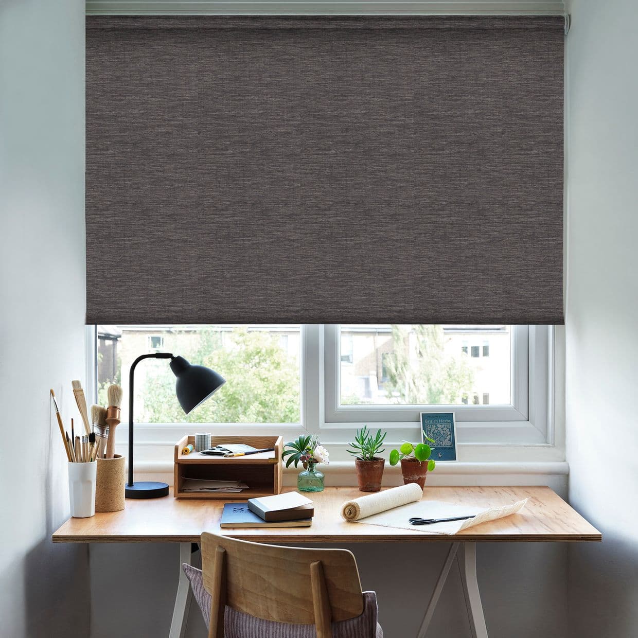 Dark gray textured roller blind lowered nearly to the sill, blocking light; above a wooden desk with a lamp, art brushes, notebooks, small potted plants, and a daylight view outside.

BRITISH BIRDS