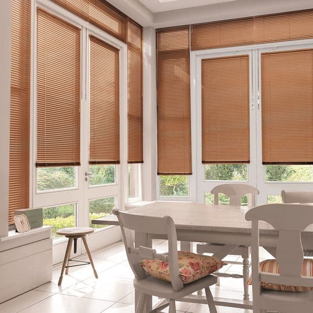 Brown horizontal Venetian blinds cover tall French-style windows and doors, lowered with slats mostly closed, filtering soft daylight into a bright dining nook with a wooden table, chairs, and garden view.