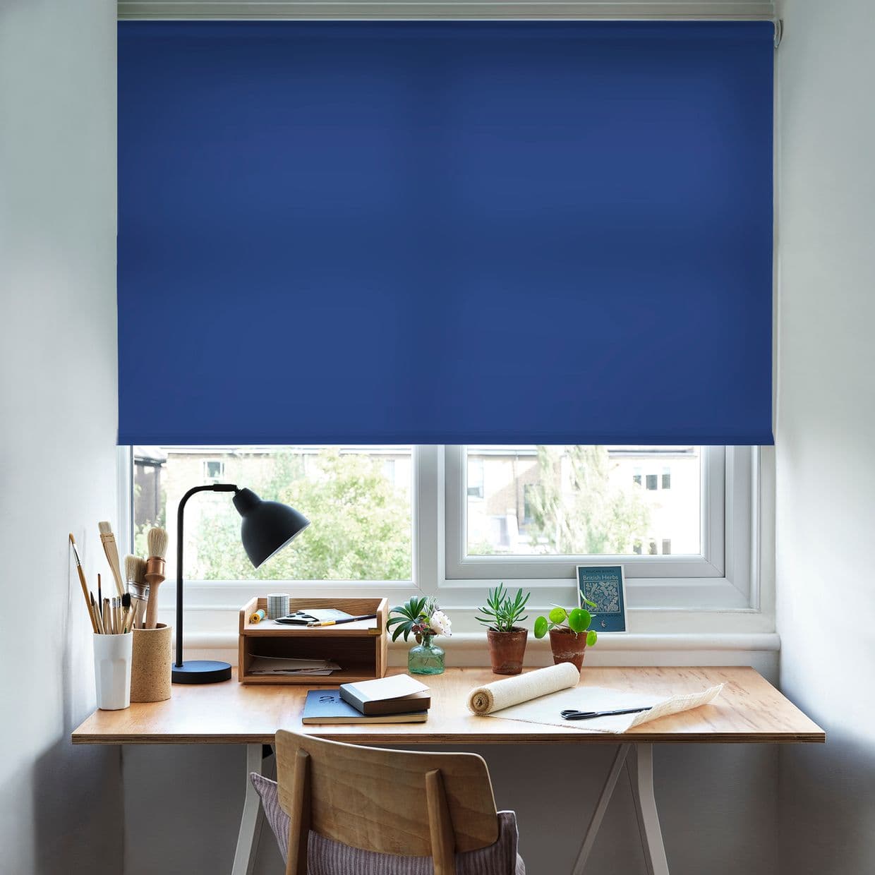 Blue roller blind, lowered across a double window, solid deep-blue fabric blocking most daylight; a sunlit home desk below with lamp, art supplies, potted succulents and an outside view.

British Herbs