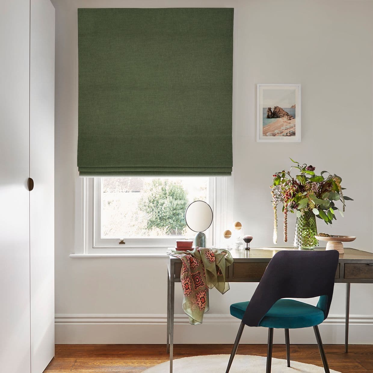 Green fabric Roman blind lowered to just above the sill, soft folded hem filtering daylight; small home office with a wooden desk, teal-upholstered chair, vase of greenery, mirror and framed seaside print.
