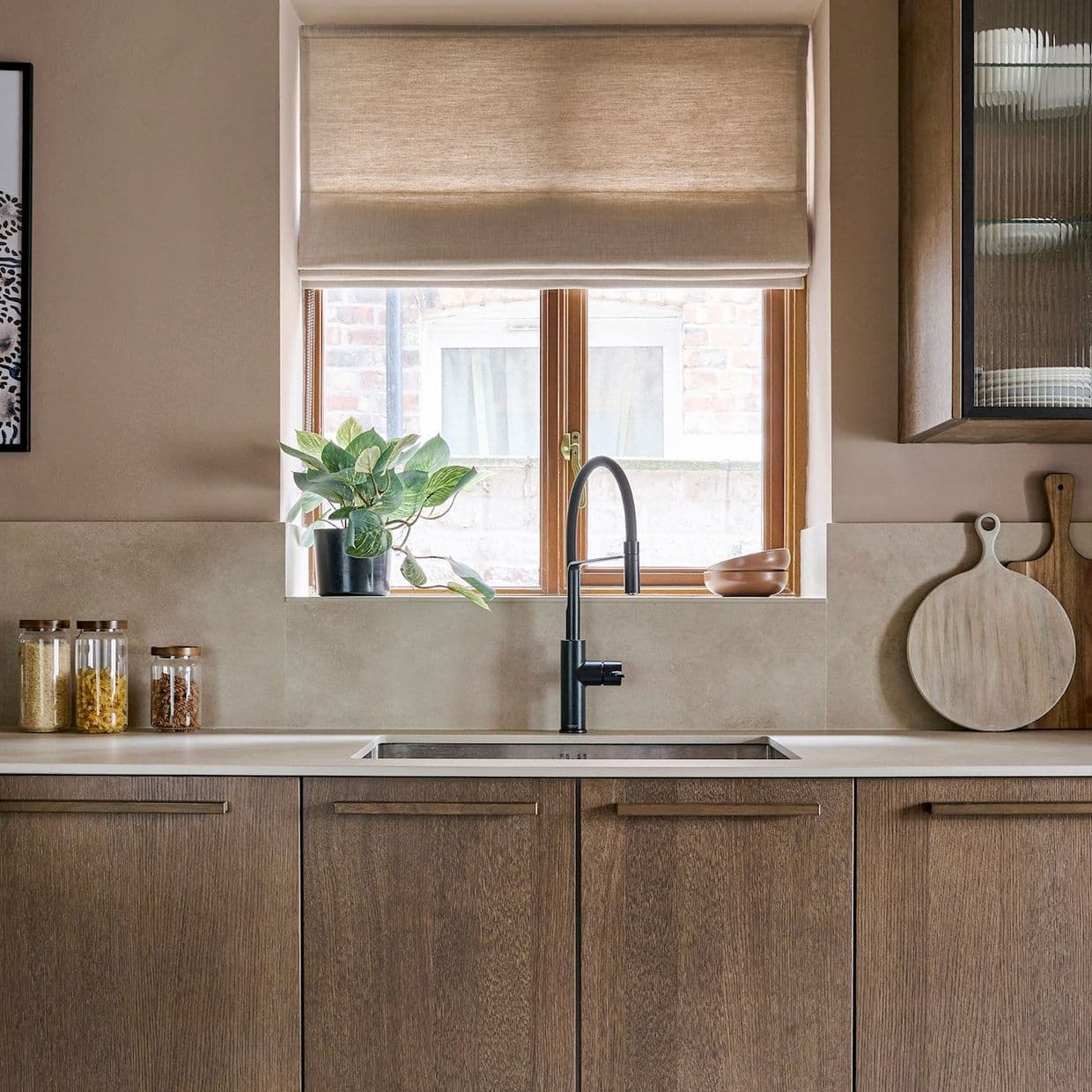 Beige fabric Roman shade, partially lowered and filtering soft daylight; framing a wooden-framed window above a kitchen sink with black faucet, potted plant on the sill, and neutral-toned wood cabinets.