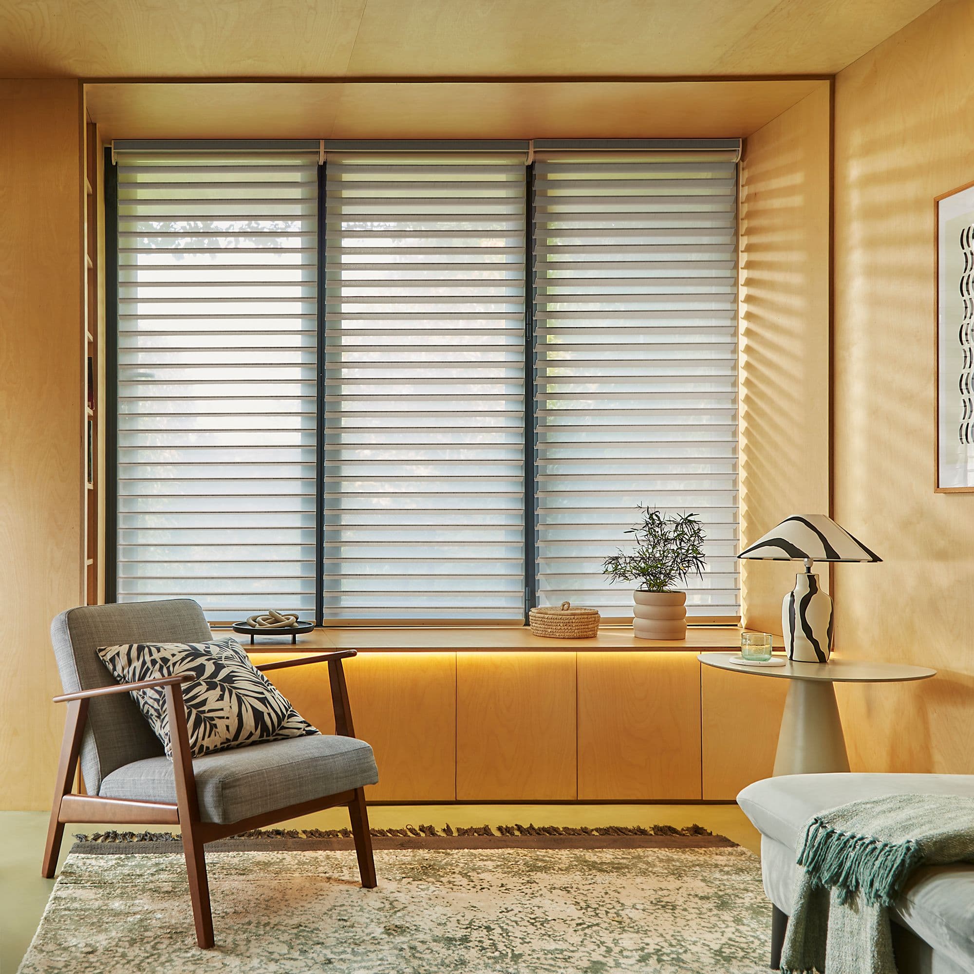 Layered horizontal roller shades across a three-panel window, evenly lowered with alternating sheer and opaque stripes, filtering soft daylight into a warm woodâ€‘paneled sitting alcove with armchair and potted plant.