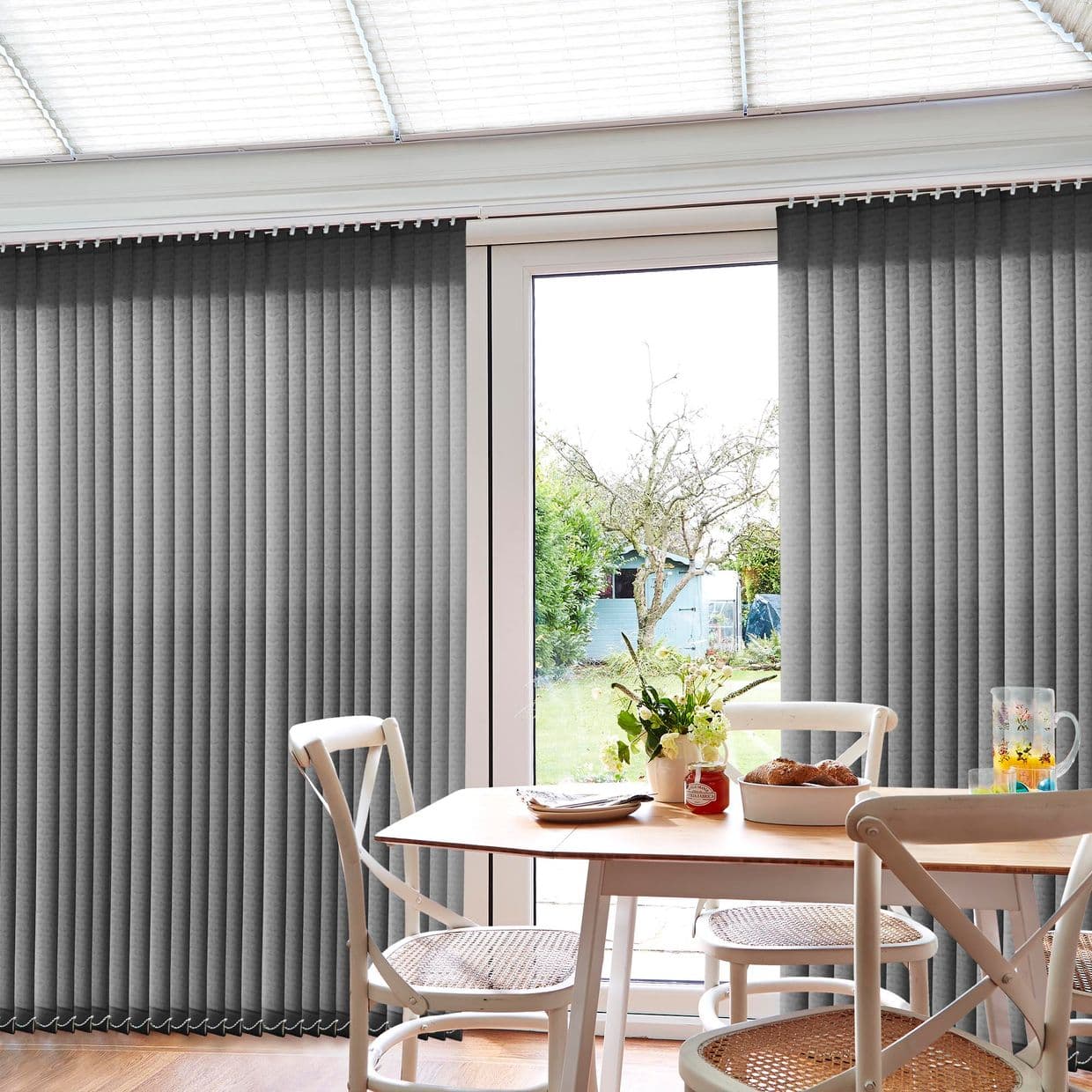 Grey vertical blinds on a track, mostly closed with a central gap; blocking light while revealing a glass door; small dining area with table, chairs, vase, and garden beyond.