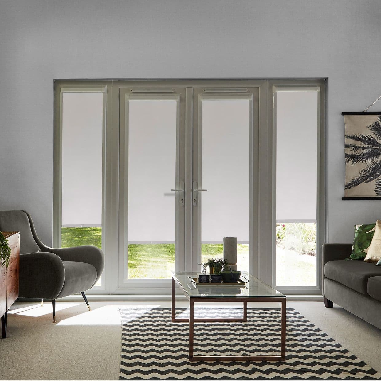 White roller blinds on four-panel French doors and sidelights, lowered leaving narrow bottom strips; filtering light and showing lawn; modern living room with grey armchair, sofa and glass coffee table.