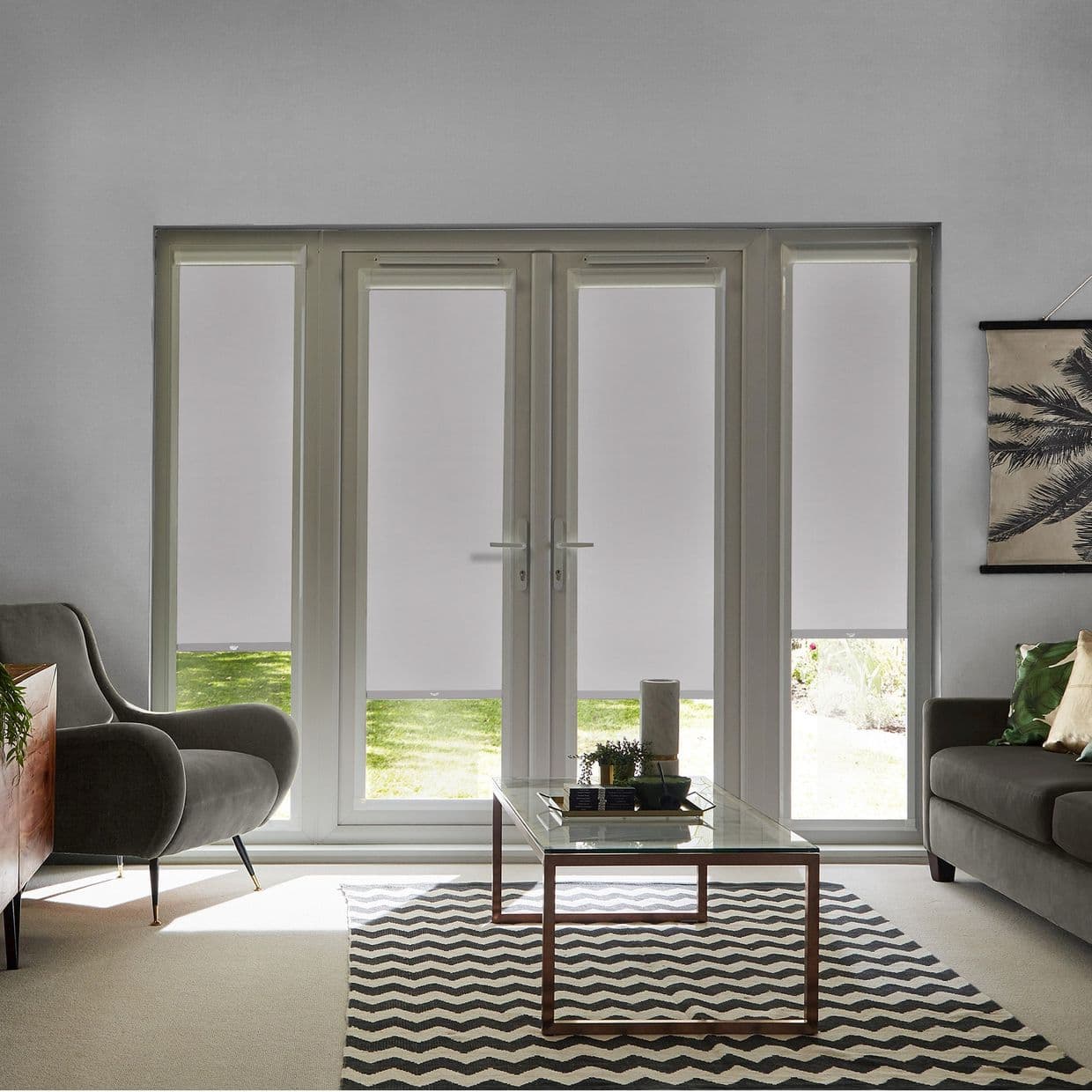 White roller blinds cover four tall glazed panels (central doors and sidelights), lowered leaving narrow bottom gaps; filtering light and obscuring view; living room with armchairs, coffee table, rug, lawn.