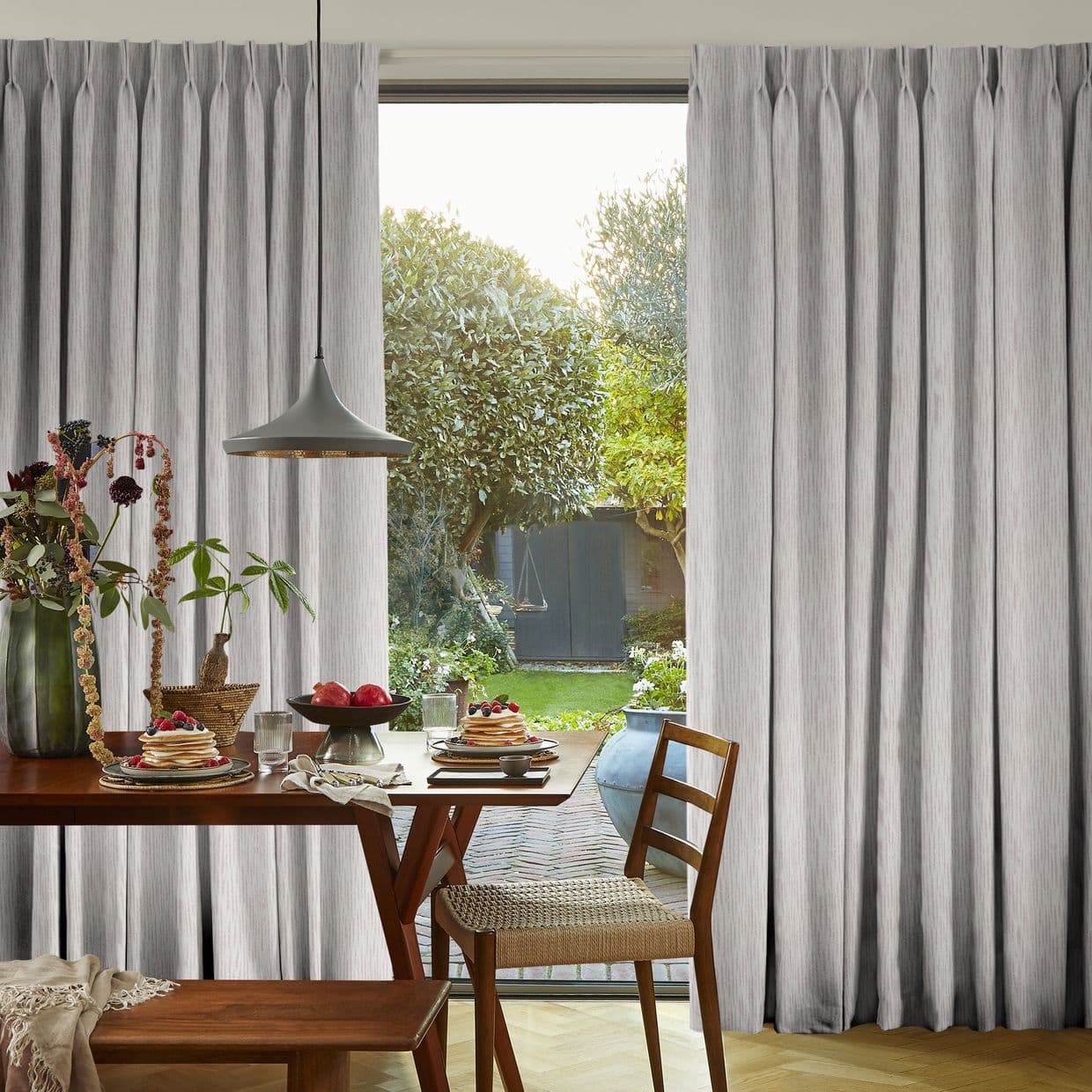 Floor-to-ceiling pleated grey curtains, gathered at the sides, partially open to frame a sunlit garden; dining table with place settings, pendant lamp and wooden chairs in the foreground.