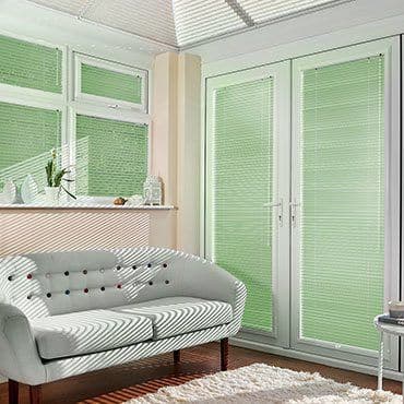 Green pleated blinds cover the windows and French doors, closed and filtering sunlight, casting striped shadows across a buttoned loveseat in a bright living room with plants on the sill.
