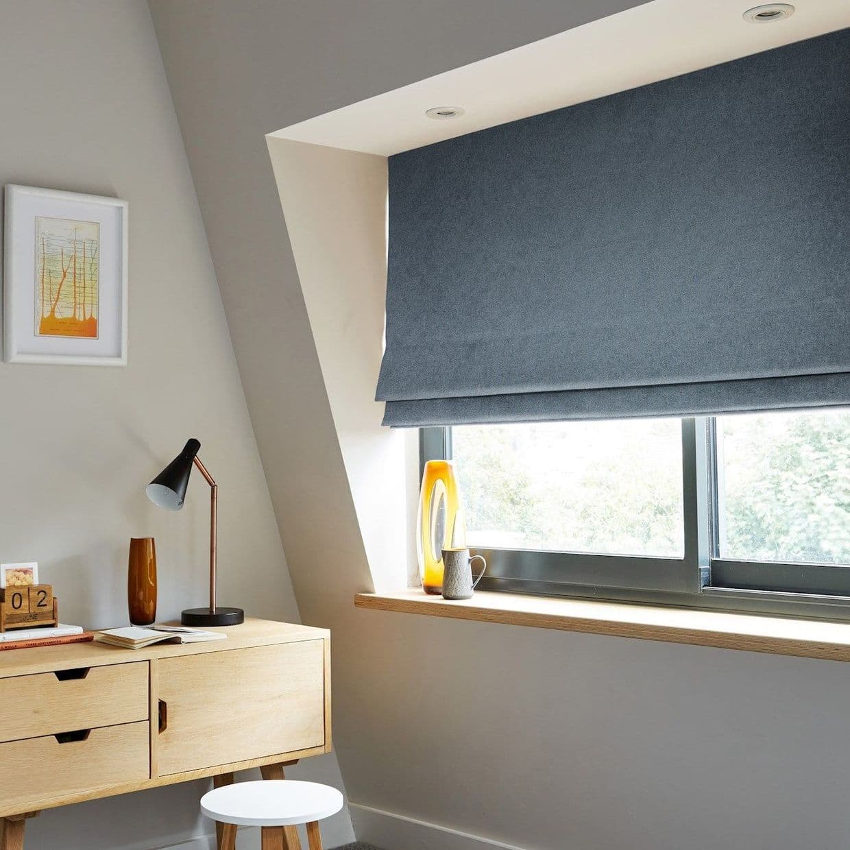 Blue Roman Roman blind lowered in layered folds, covering the upper casement window and partially filtering daylight; small slopedâ€‘ceiling home office with light wood desk, lamp, stool, vase on sill. 02 JUNE