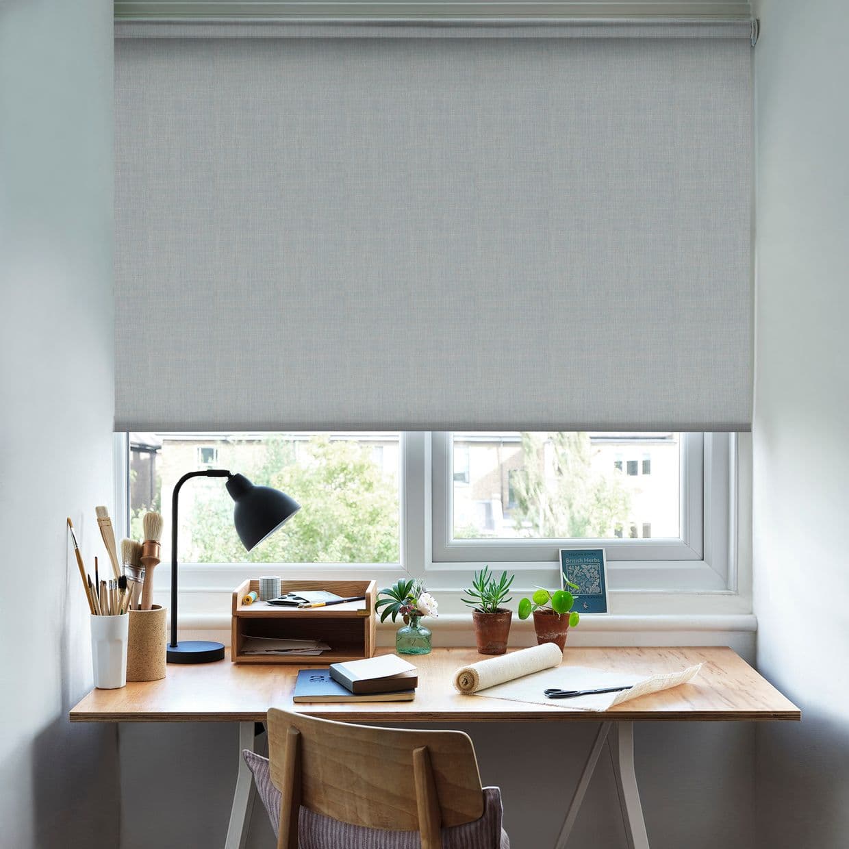 Grey fabric roller blind, lowered over a double window, filtering soft daylight; a wooden desk beneath holds a black lamp, paintbrushes, notebooks, and small potted succulents in a home workspace.
