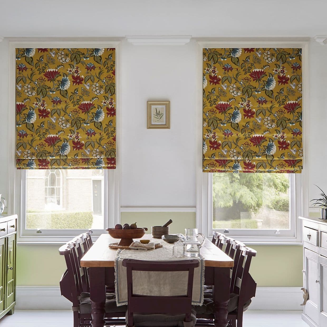 Two mustard-yellow floral Roman blinds, lowered over adjacent windows, patterned with red and blue flowers, filtering daylight into a dining room with wooden table, chairs and garden view.