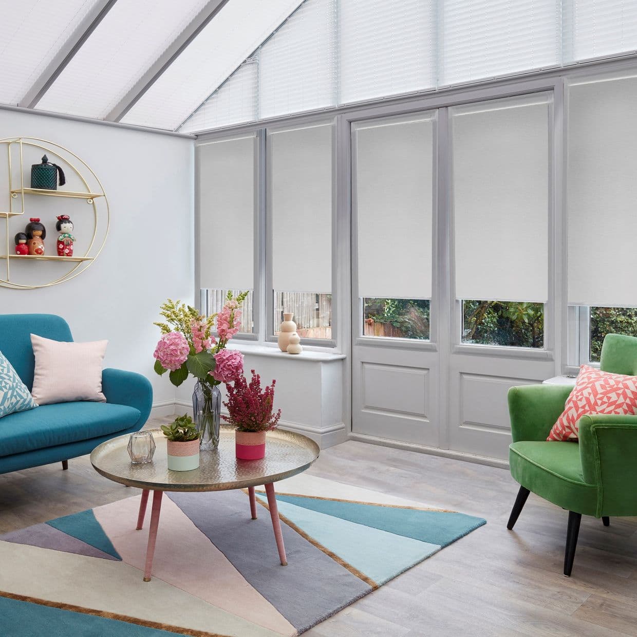 Light-gray roller blinds covering four tall glazed doors and triangular roof windows, lowered to mid-height and filtering daylight; bright conservatory with teal and green armchairs, round coffee table, pastel rug, plants.