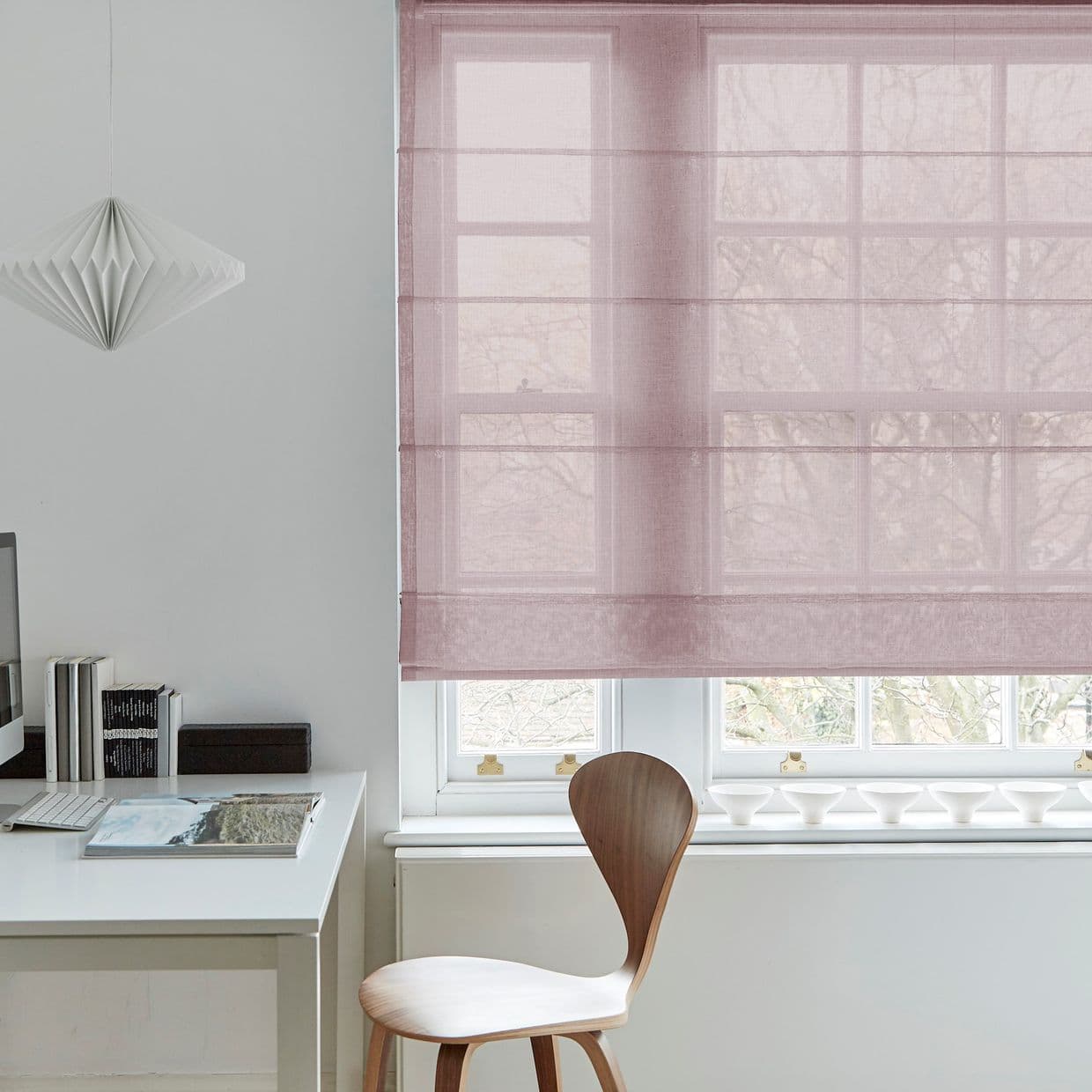 Sheer mauve Roman blind, lowered and softly filtering daylight; wooden chair and white desk beside window, pendant paper lamp overhead, minimalist home office with pale walls and bare tree view.