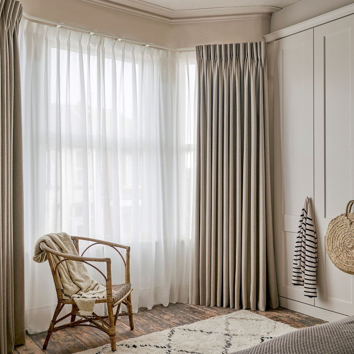 Floor-to-ceiling pleated beige curtains with sheer white voile panels across a corner bay window, filtering soft daylight into a neutral, cozy bedroom with rattan chair, wooden floors, rug and wardrobe.