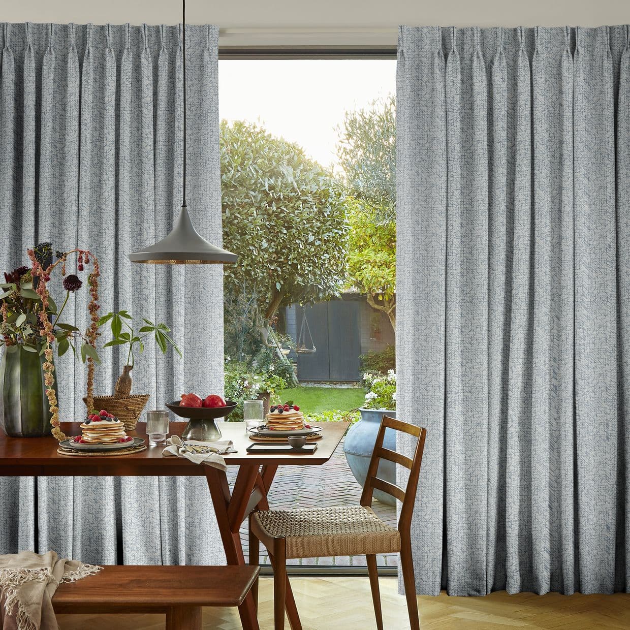 Pleated grey full-length curtains, drawn aside to frame the window opening and filter daylight, in a dining room with a set table, pendant lamp and a sunlit garden visible beyond.