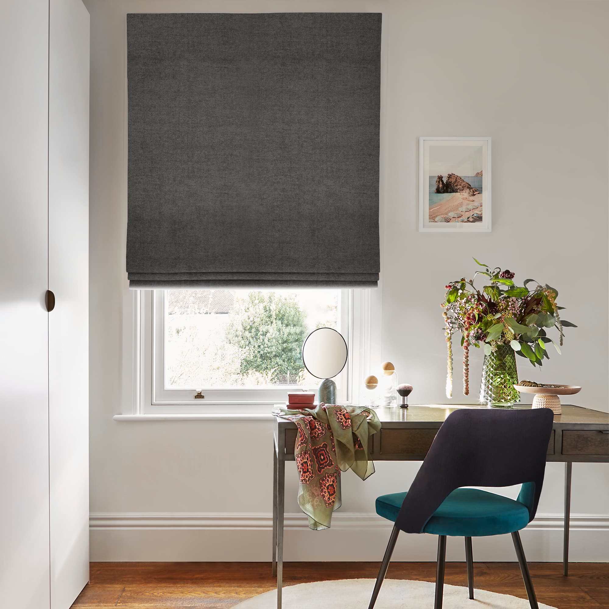 Dark grey Roman blind lowered over window, mostly closed and filtering daylight with an open strip at bottom; above desk with mirror, teal chair, floral vase in bright room.