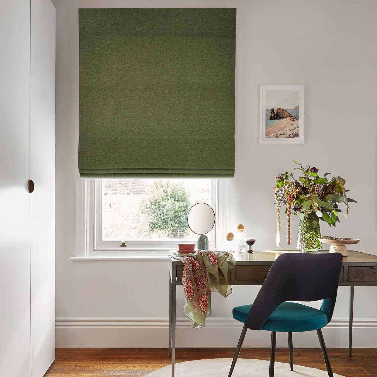 Olive-green fabric Roman blind, fully lowered with neat horizontal folds, filtering soft daylight through a narrow bottom gap; compact home workspace with desk, teal chair, mirror and vase of flowers.