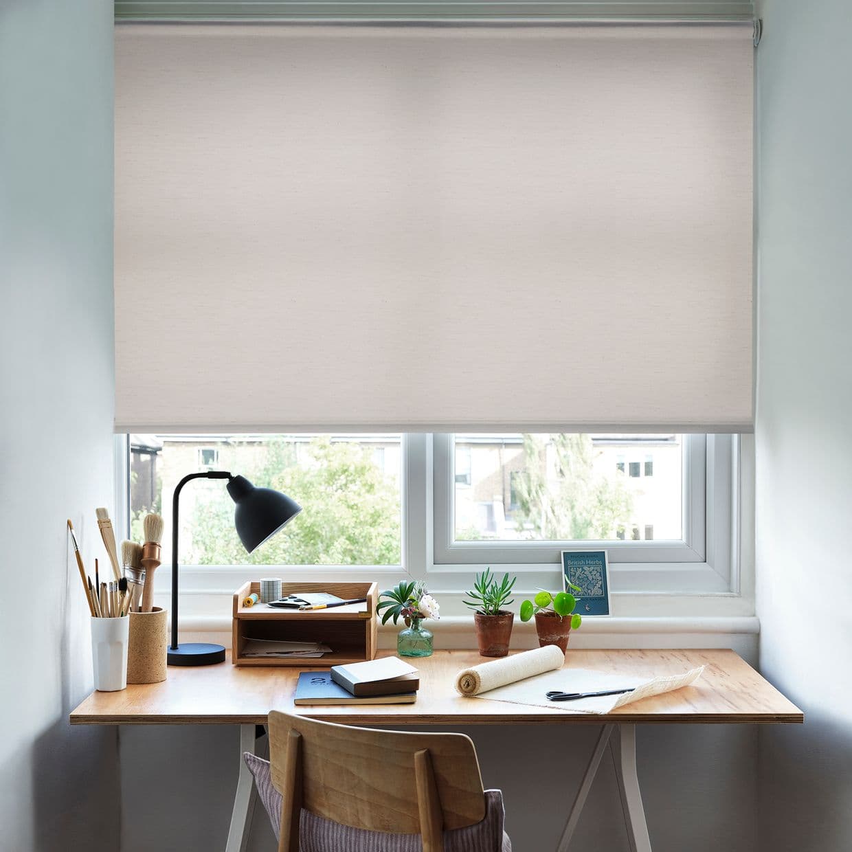 Light beige roller blind, lowered three-quarters and mounted inside the frame, filtering soft daylight over a tidy home desk with lamp, paintbrushes, potted succulents, notebooks, and a leafy outside view.