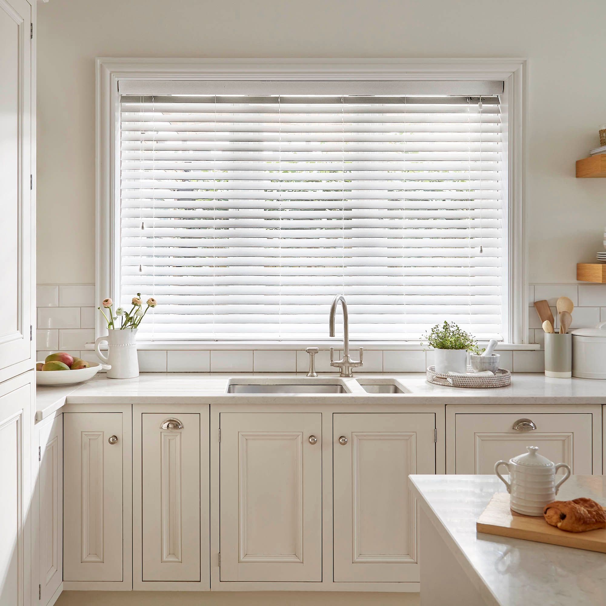 White horizontal Venetian blinds covering a large kitchen window, lowered and slightly tilted to diffuse daylight; stainless-steel faucet and sink beneath, light-filled neutral-toned kitchen with countertop plants and utensils.
