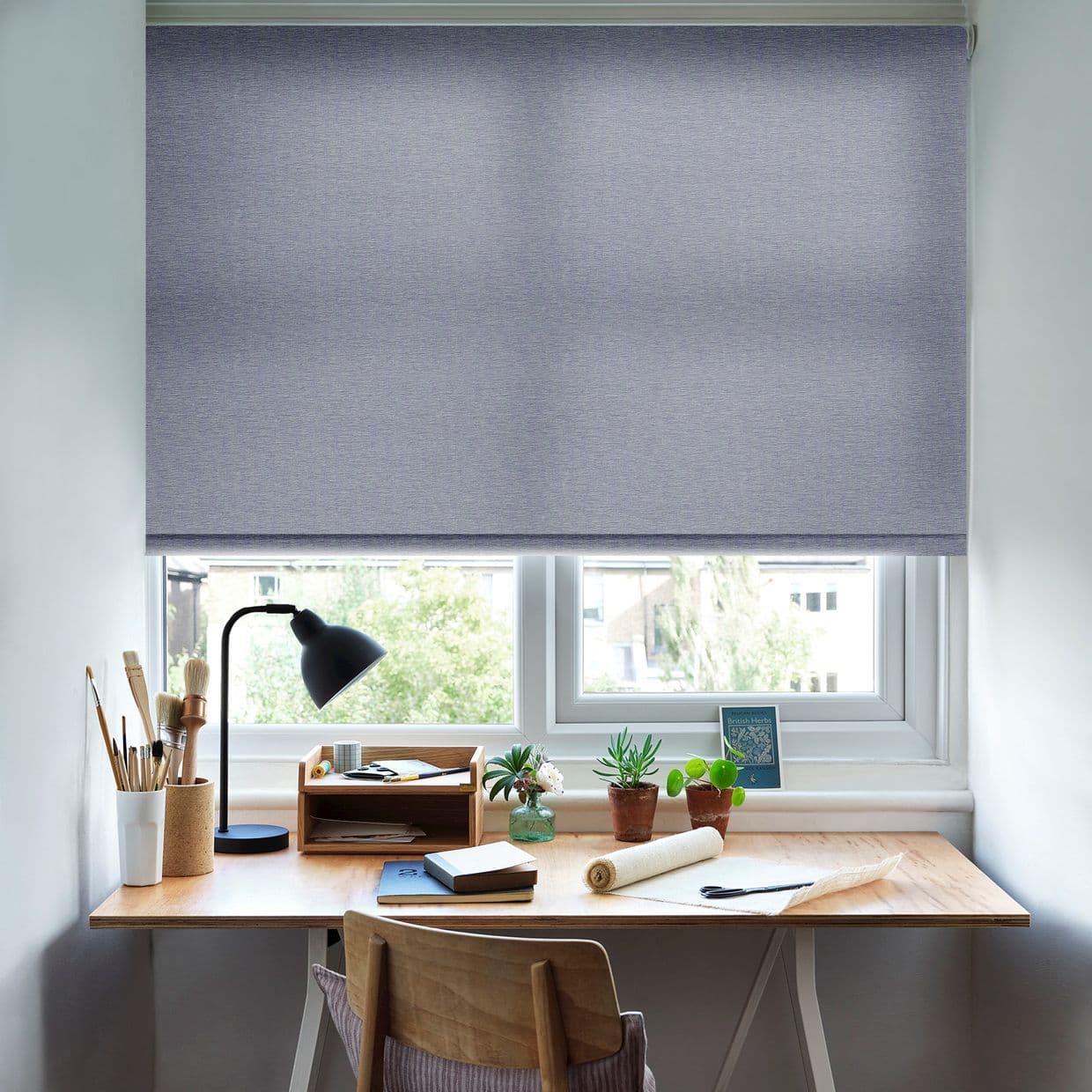 A grey roller blind, lowered across the window with a slim bottom gap, filtering daylight. It shades a tidy wooden desk with lamp, brushes, notebooks and small potted plants.