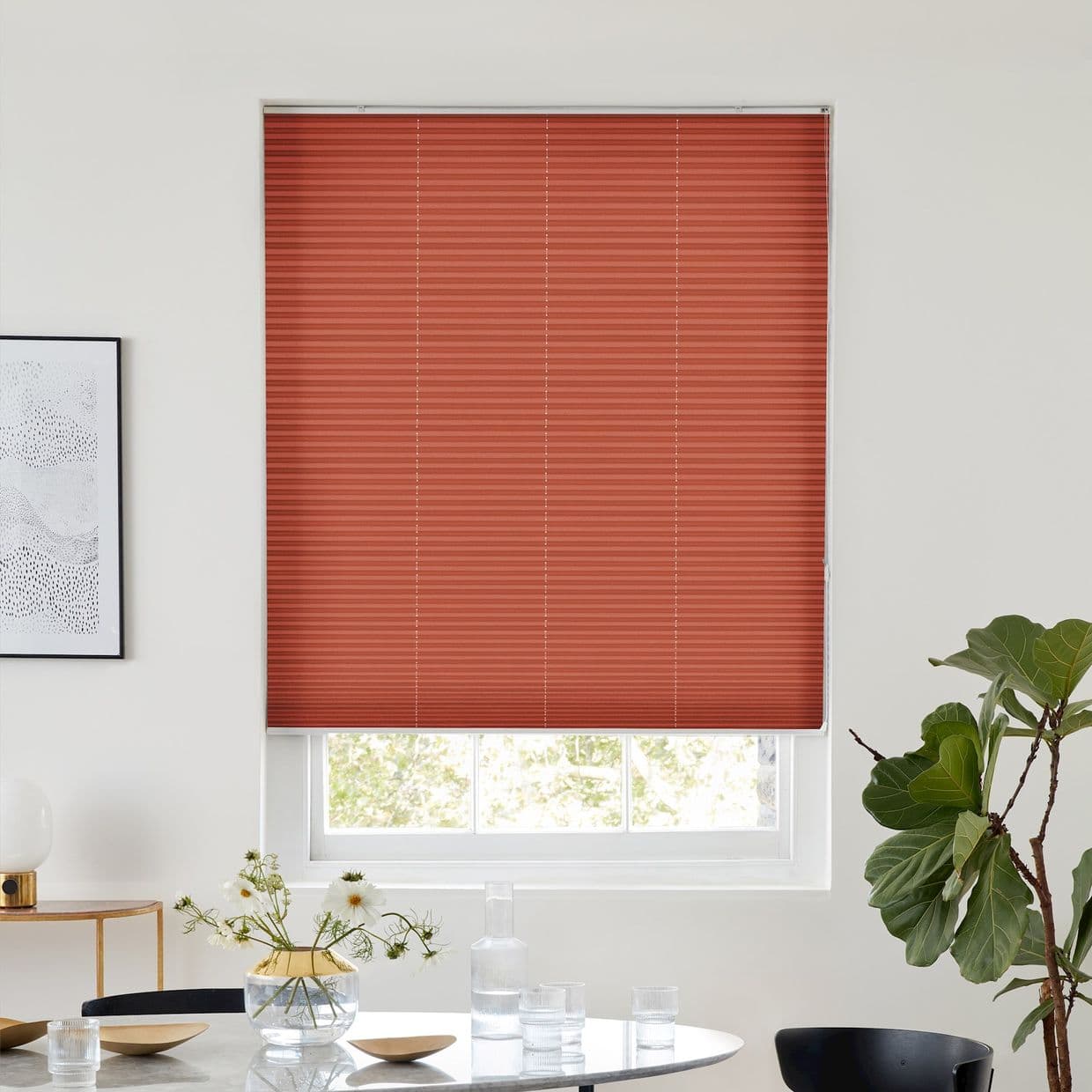 Red pleated blind, lowered over the window with a narrow bottom gap, filtering daylight; in bright dining space with table, vase of flowers, side lamp and fiddleâ€‘leaf fig.