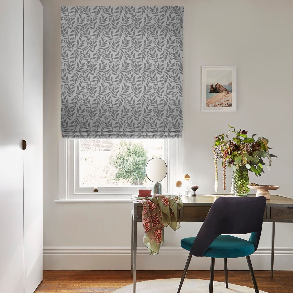 Patterned grey leaf Roman blind, lowered over the window and filtering daylight; a desk with mirror, vase of flowers and teal chair sits beneath in a neutral-toned home study.