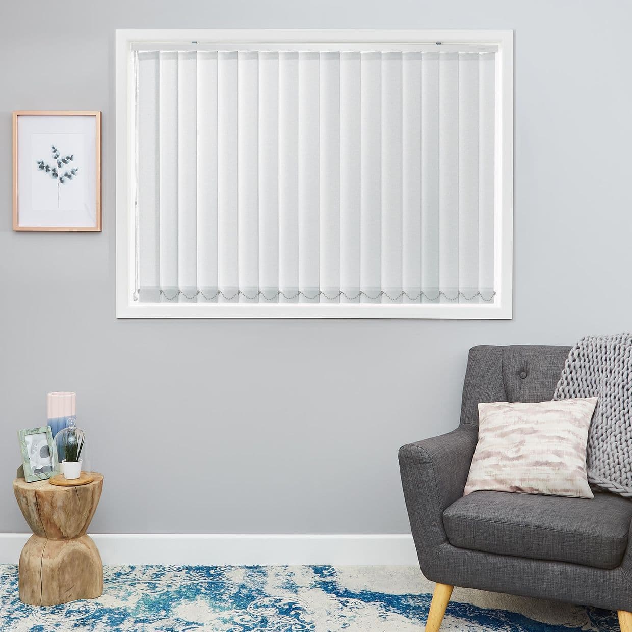 White sheer vertical blinds, fully closed with chain weights along bottoms, diffusing soft daylight; in a gray-walled living room with a gray armchair, side table and blue patterned rug.
