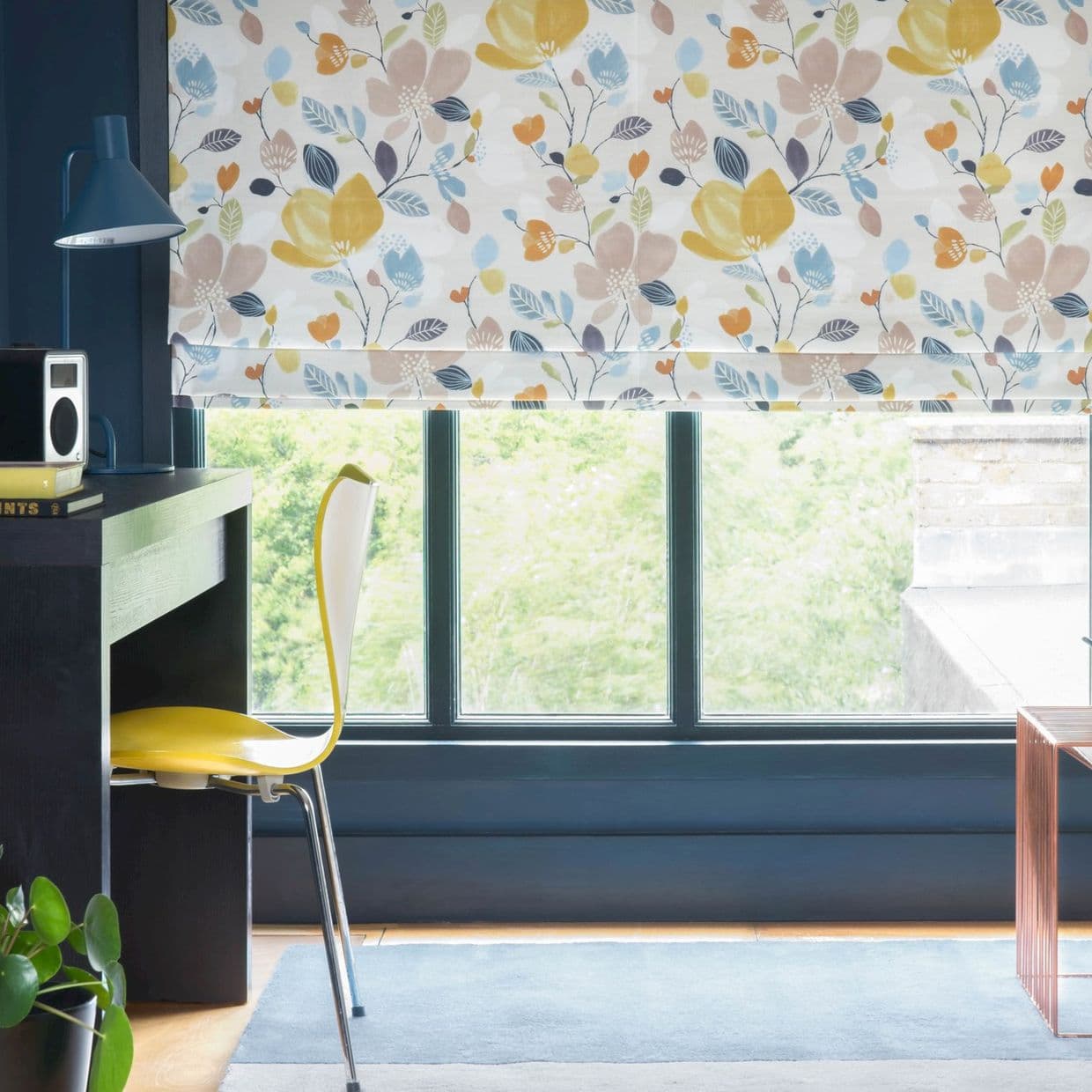 Floral-patterned Roman blind lowered across a three-panel window, filtering daylight; a dark-painted desk with blue lamp and yellow chair sits beside window in a bright home office overlooking trees.