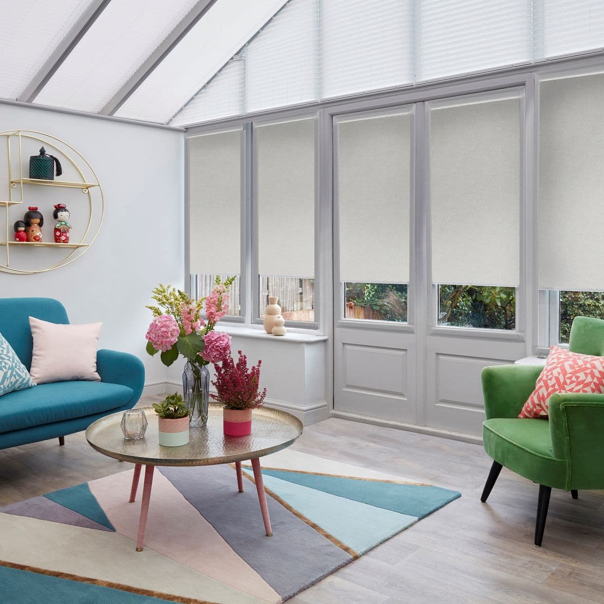 Beige roller blinds, lowered over upper panes and partially over lower panes, filtering soft daylight; in a bright conservatory-style living room with slanted glass roof, colourful sofas, rug, and flowers.