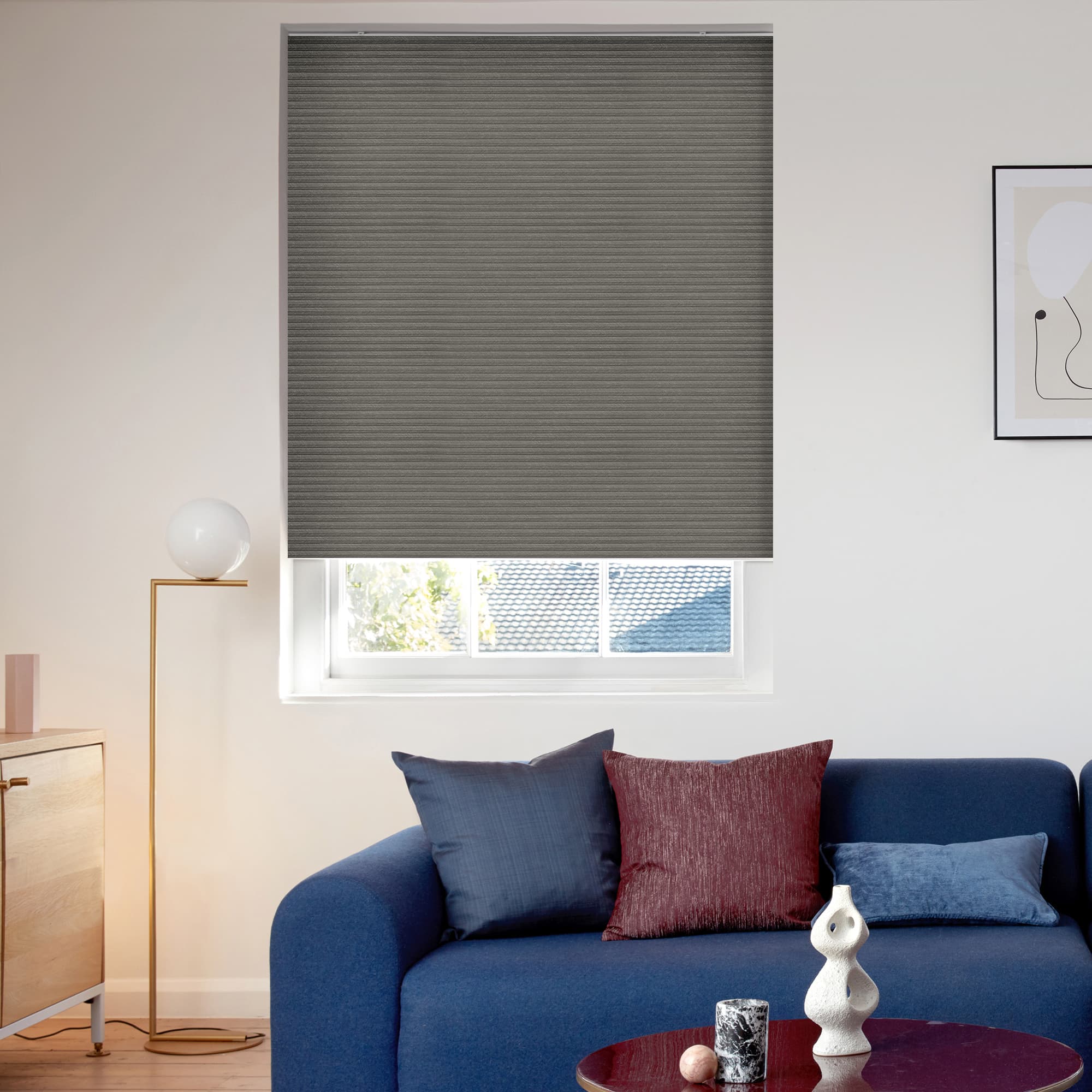 Gray pleated cellular blind, fully lowered over the window with subtle horizontal texture; closed and filtering daylight, set in a bright living room above a blue sofa, lamp, and artwork.