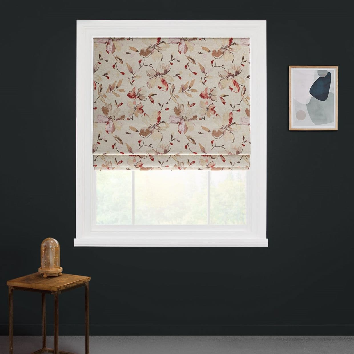 Floral Roman blind (beige with pink-brown leaves), partially lowered and filtering daylight, covers a window on a dark gray wall; nearby a wooden table with glass-dome lamp and abstract art.