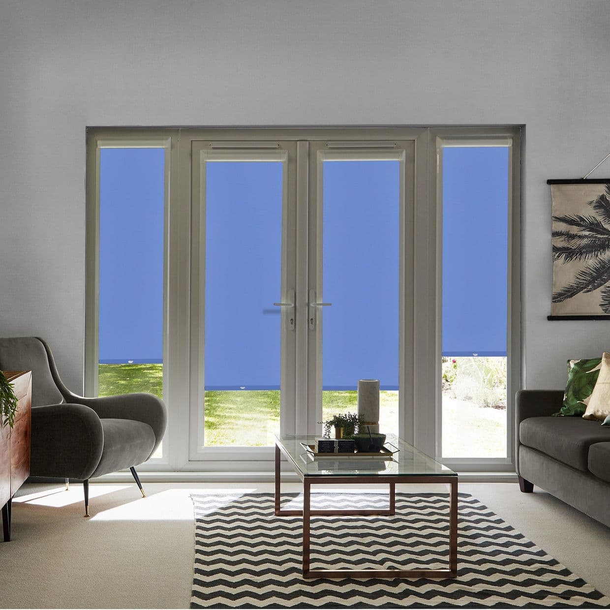 Four blue roller blinds (narrow side panels and double French doors), fully lowered with small bottom gaps, blocking view and filtering light; modern living room with sofas, glass coffee table, garden outside.