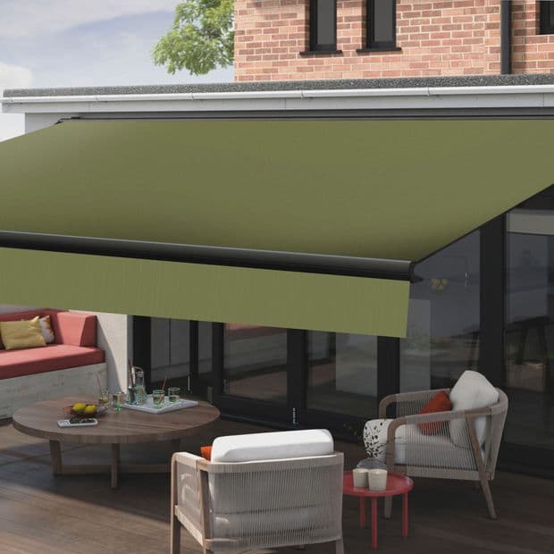 Green retractable awning with black metal bar and arms, fully extended above sliding glass doors, shading a sunlit patio with wicker chairs, round table, built-in bench and brick wall.