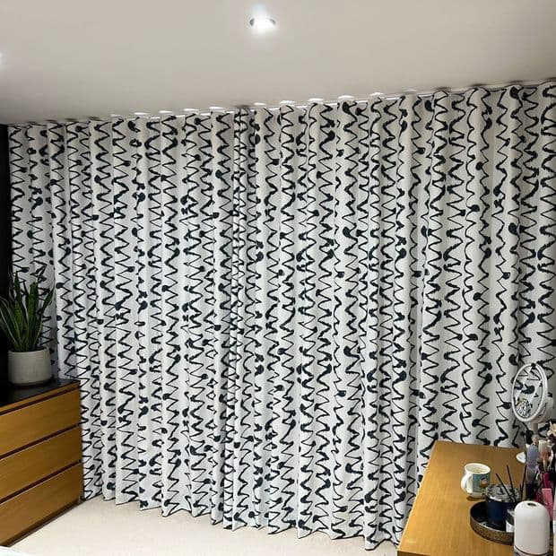 Full-length white fabric curtains with irregular black squiggle pattern, hanging from a ceiling track and drawn closed, filtering light; room corner with wooden dresser, potted plant, desk, ceiling light.