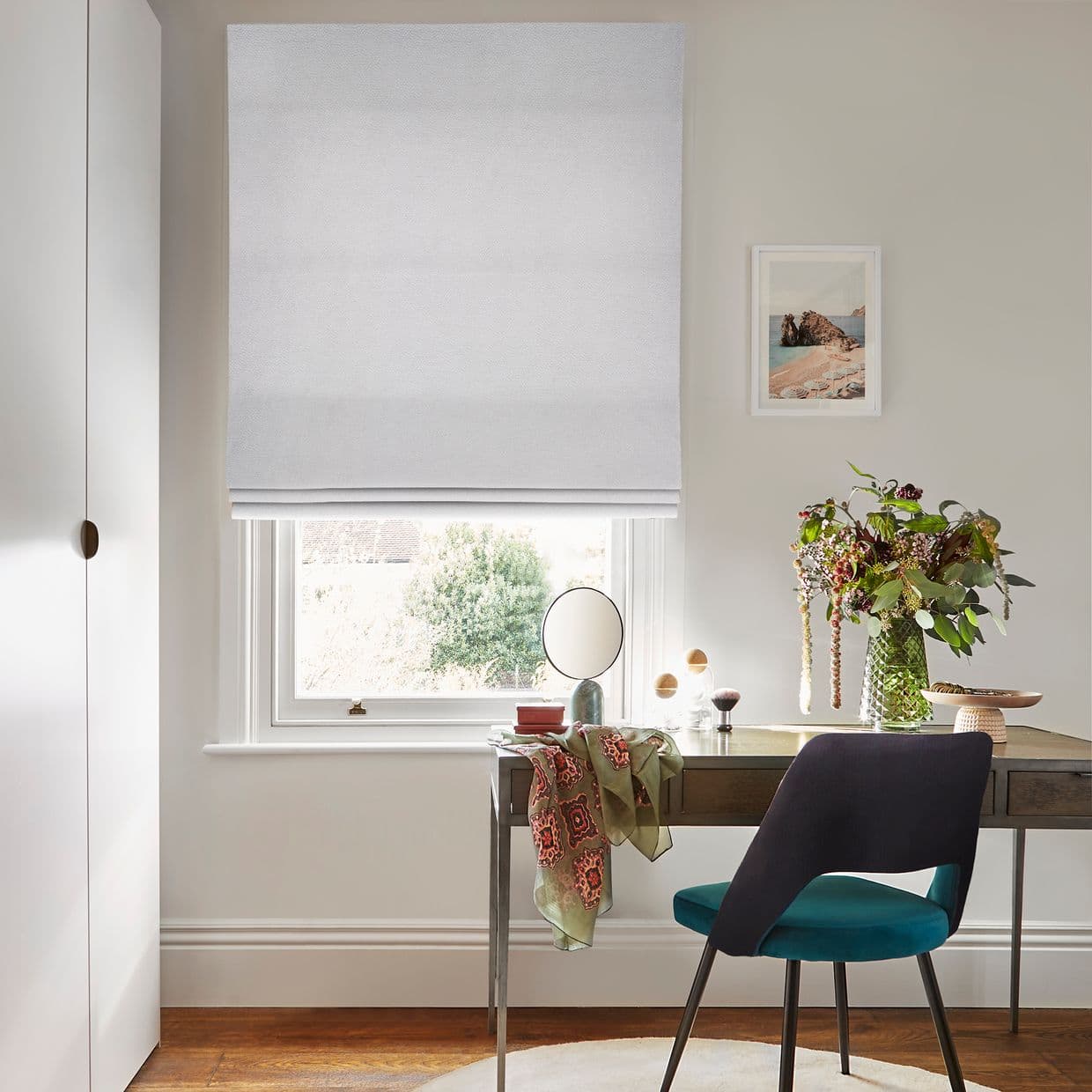White fabric Roman blind, lowered with a soft fold, filtering daylight; a desk with vase of flowers and a teal chair in a bright home office overlooks shrubs.