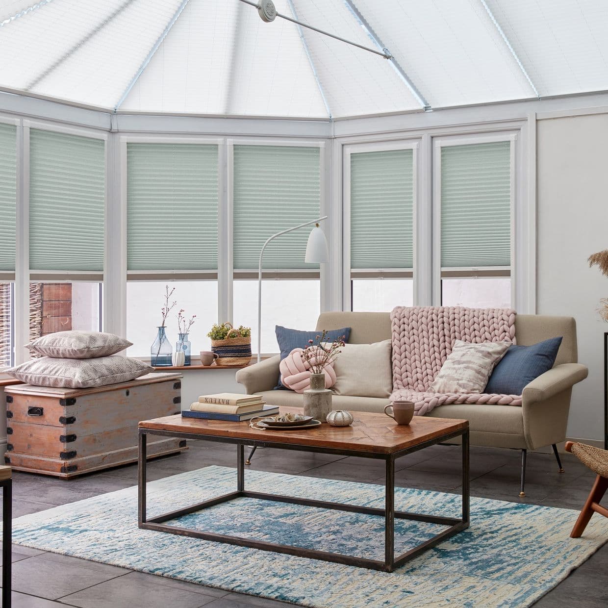 Honeycomb pleated blinds across tall conservatory windows, pale seafoam, lowered, leaving narrow bottom gap; filtering daylight. Context: bright sunroom with beige sofa, chunky pink throw, wooden coffee table and rug.