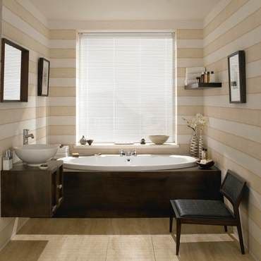 White horizontal Venetian blinds covering the central window, lowered and closed, filtering soft daylight; bathroom with dark wood-clad built-in bathtub, vessel sink, shelves, chair, striped beige walls and tiled floor.