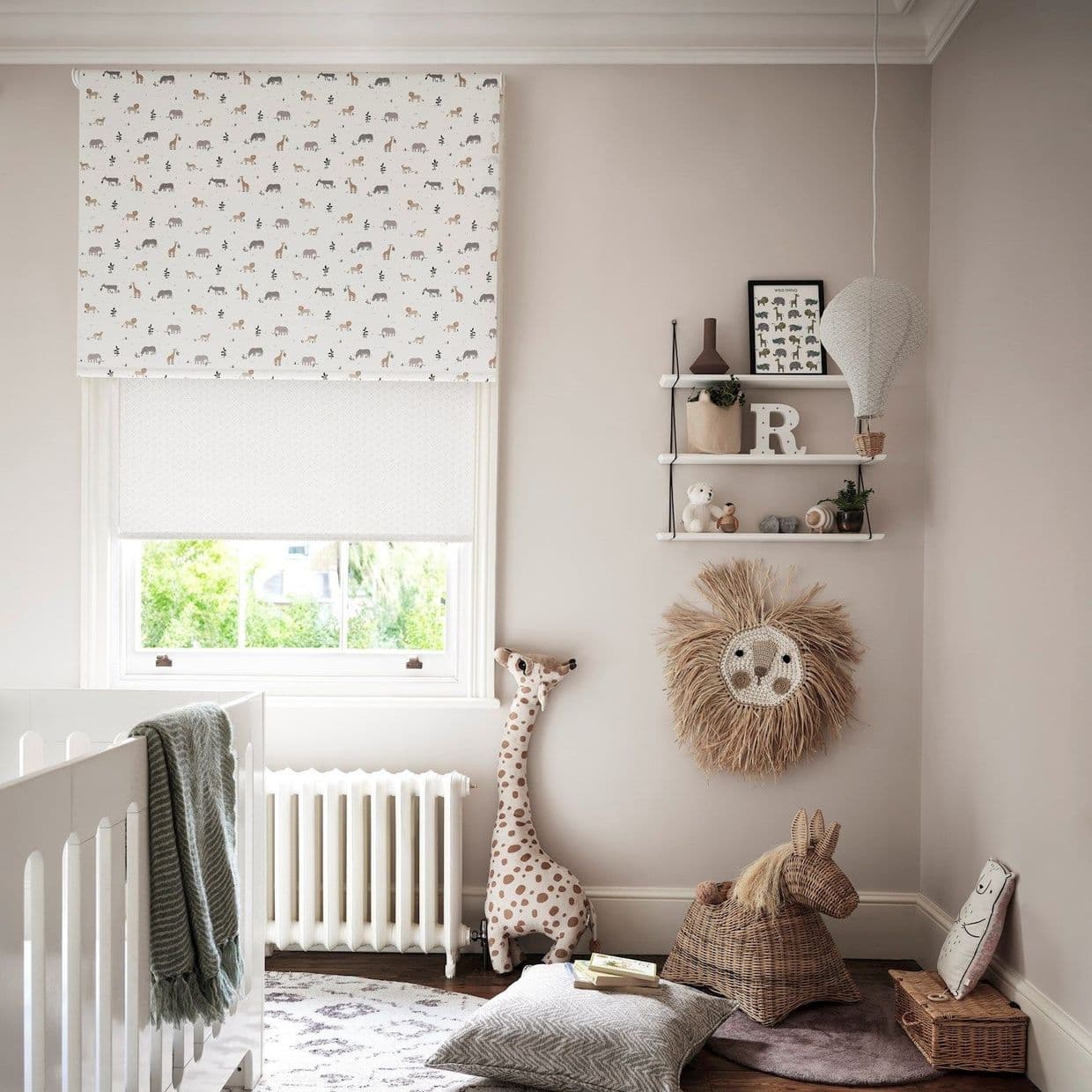 A white roller blind with small animal prints lowered over a window, filtering soft daylight; neutral nursery with crib, stuffed giraffe, wicker horse, shelves, hanging hot-air balloon and woven lion wall decor.

R
