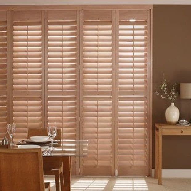 Wooden plantation shutters cover floor-to-ceiling doors, closed with tilted slats filtering sunlight and casting striped shadows; warm dining area visible with glass table, place settings and side table with vase.
