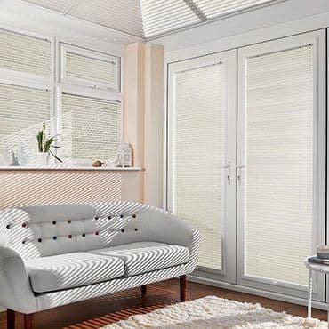 White pleated blinds fitted inside French doors and adjacent windows, lightâ€‘filtering; closed, softening daylight and casting striped shadows across a pale sofa and wood floor in a sunlit living room.