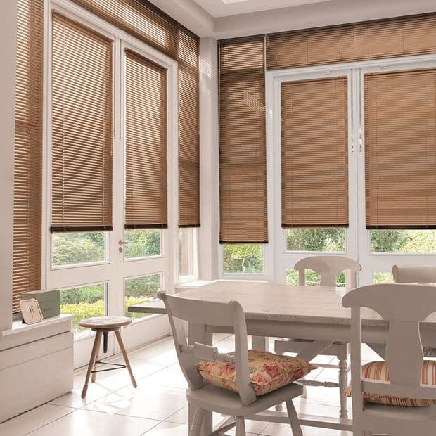 Brown horizontal Venetian blinds cover tall windows and patio doors, lowered and mostly closed, filtering soft daylight into a sunlit dining nook with white table and chairs, floral cushions, garden glimpses.