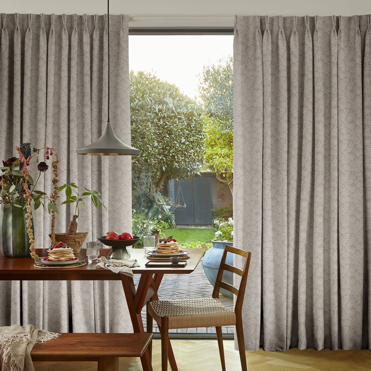 Full-length pleated patterned beige curtains frame a sliding glass door, partially drawn to reveal a sunlit garden; dining table set for breakfast with pancakes, chairs, and a hanging pendant light.
