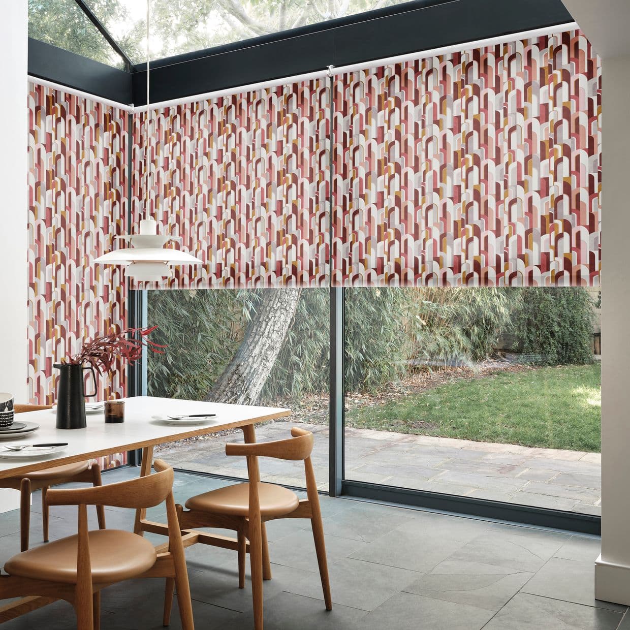 Patterned roller blinds with multicolored geometric arch print, mounted across the upper glazing; partially lowered, filtering light while leaving lower glass visible; dining area with wooden chairs and garden view.