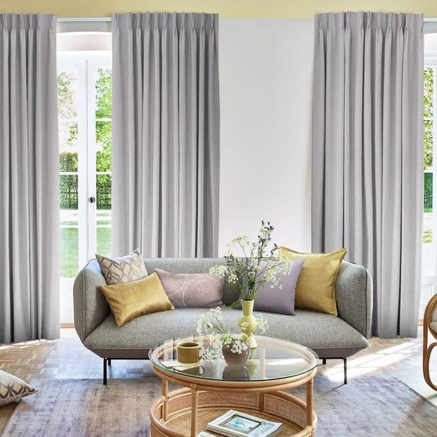 Floor-length pleated gray curtains drawn closed over French doors and flanking windows, filtering daylight into a bright living room with a curved sofa, decorative pillows and a glass coffee table.