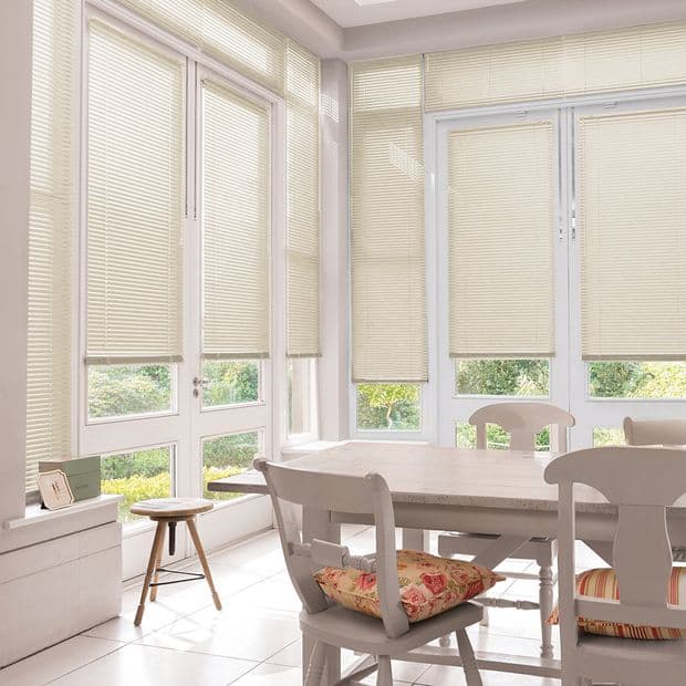 Object â€” Cream pleated blinds on tall windows and French doors. Action â€” Mostly lowered, filtering daylight through small gaps. Context â€” Bright dining nook with white table, chairs, floral cushions, garden view.