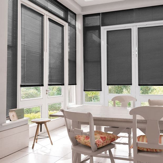 Black horizontal Venetian blinds, lowered across bay windows with narrow slats, partially blocking but letting low light and garden views through; white dining table and chairs in bright corner room.