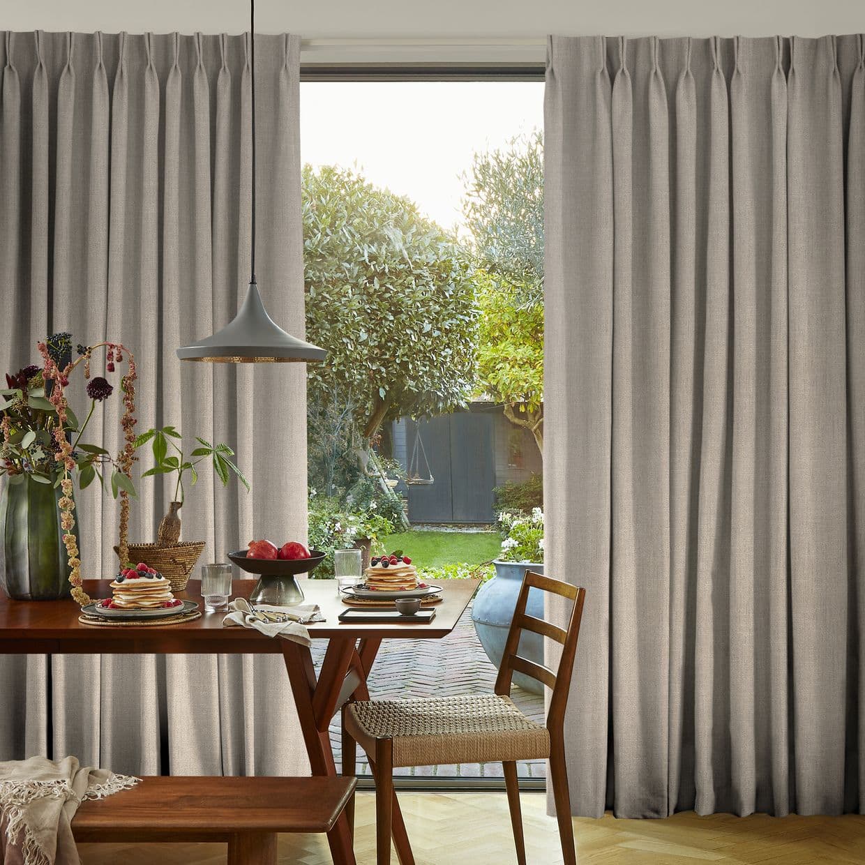 Beige full-length pleated curtains, gathered on each side and slightly parted at center revealing a sunlit garden; inside a dining table set for breakfast with wooden chairs and pendant lamp.